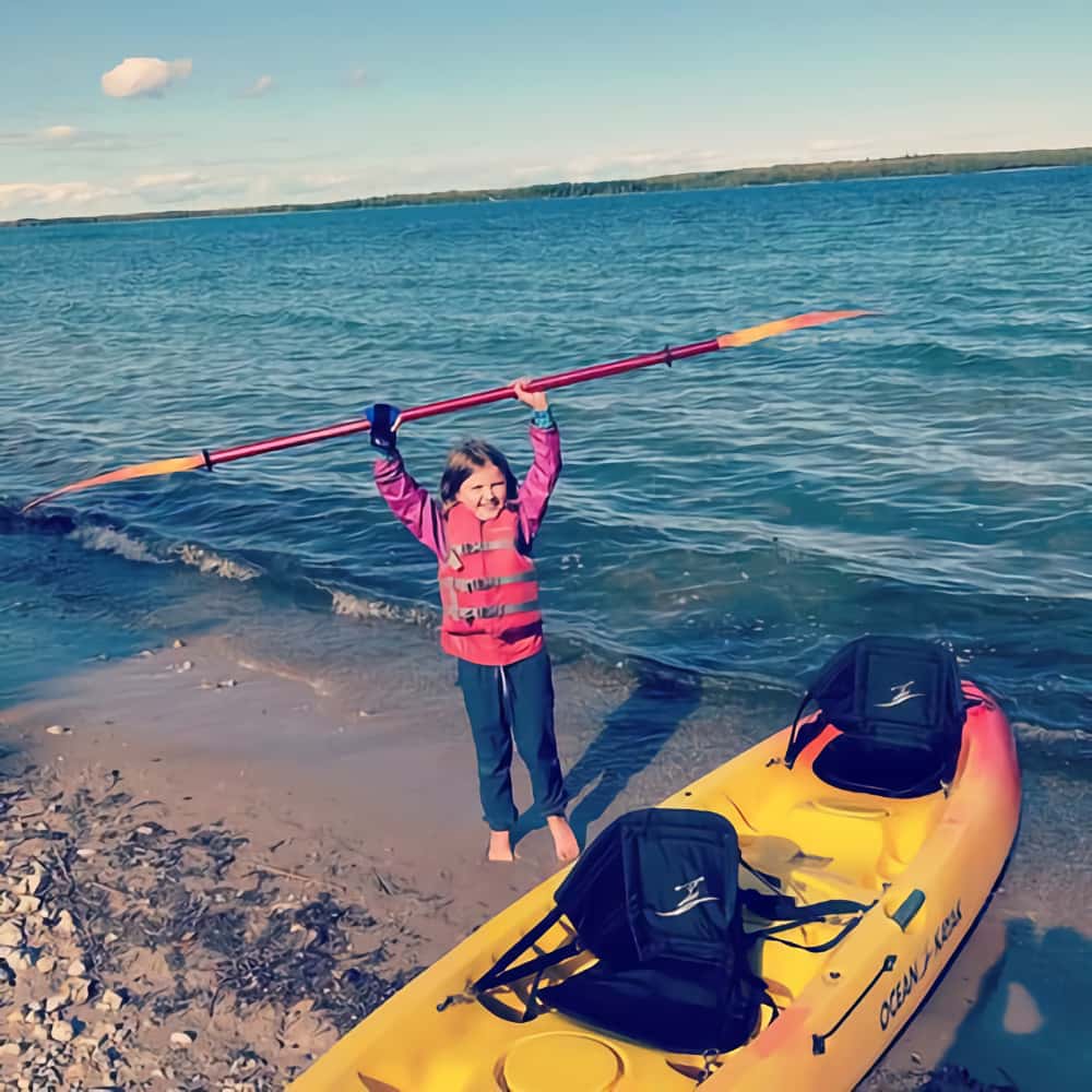 A child holding a kayak paddle over her head with a mini aid. She is by the sea and a 2-person kayak.